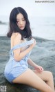 A woman in a denim dress sitting on a rock by the water.