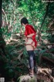 A woman in a red shirt and black stockings standing on a rock in the woods.